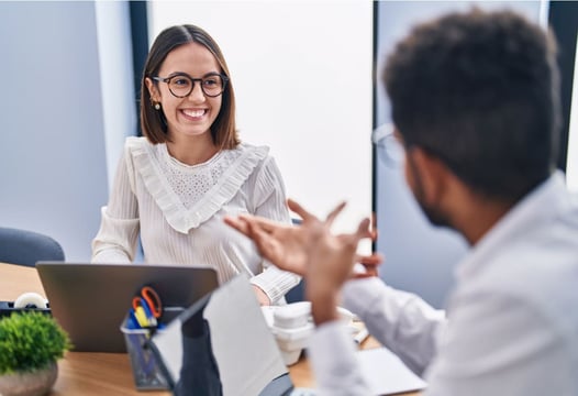 Dos personas dialogando en su oficina mientras trabajan en sus computadores portátiles.